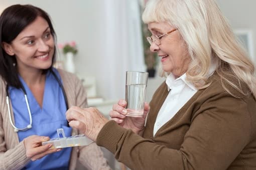 A nurse gives an older woman her pills from a pill organizer, with a glass of water